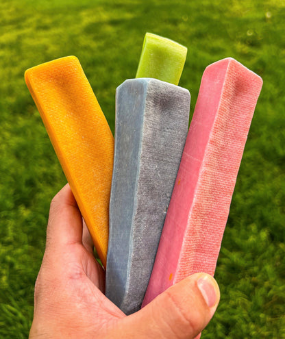 Image of a hand holding an orange, blue, green and pink yak cheese with grass in the background