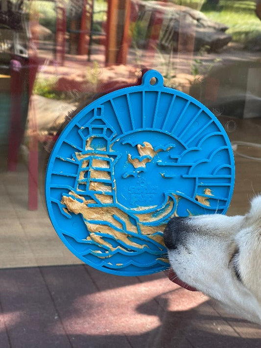 Blue circular lick mat with a lighthouse and waves pattern beside a dogs nose on a glass surface