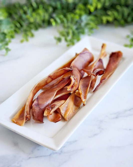 Image of multiple pig ears on a white plate on a white marble surface with greenery in the background